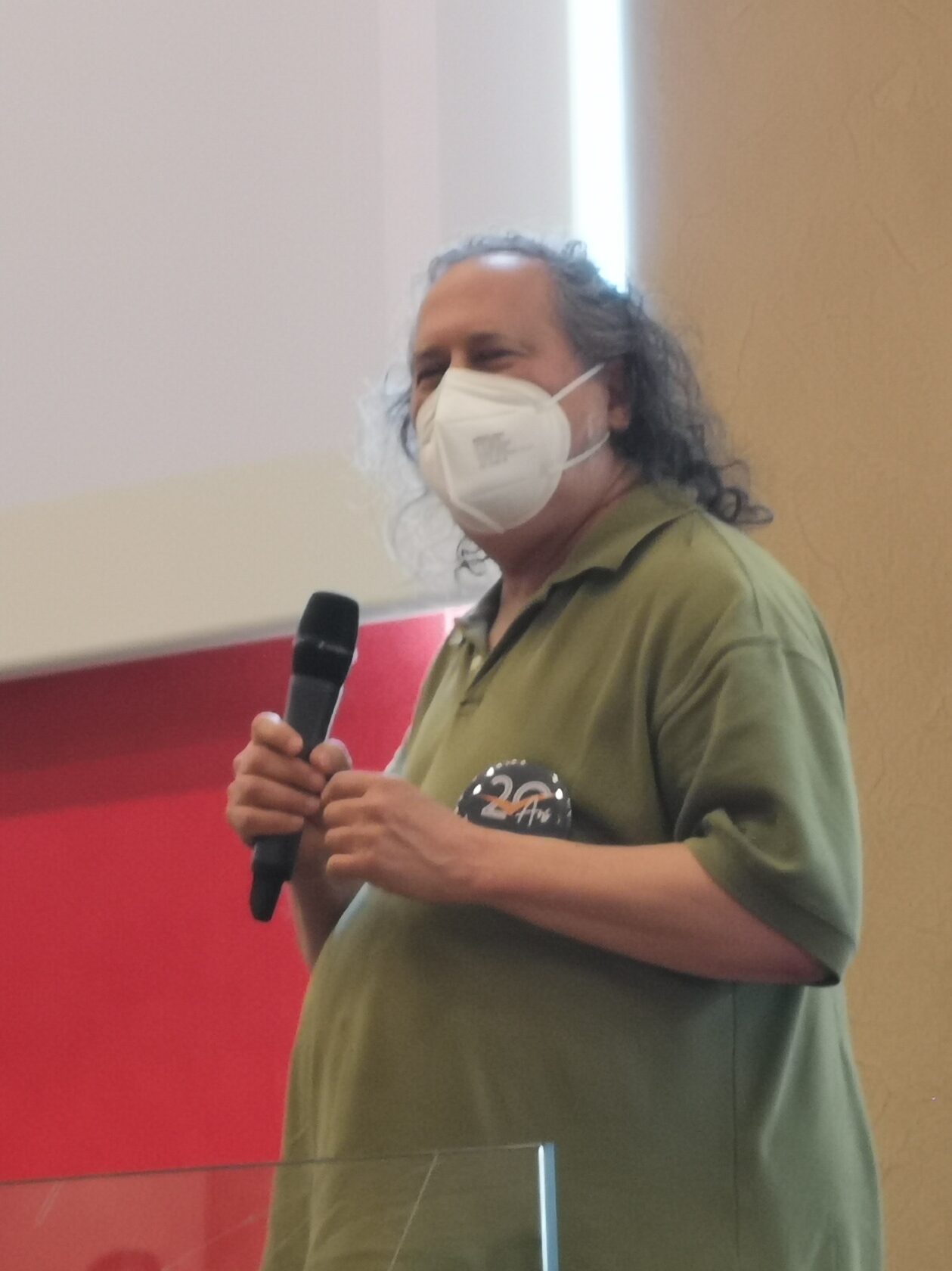 Richard M. Stallman est debout sur une estrade, il porte un micro HF à la main, un masque FFP2 devant le visage et un badge du congrès de l’adullact sur la poitrine